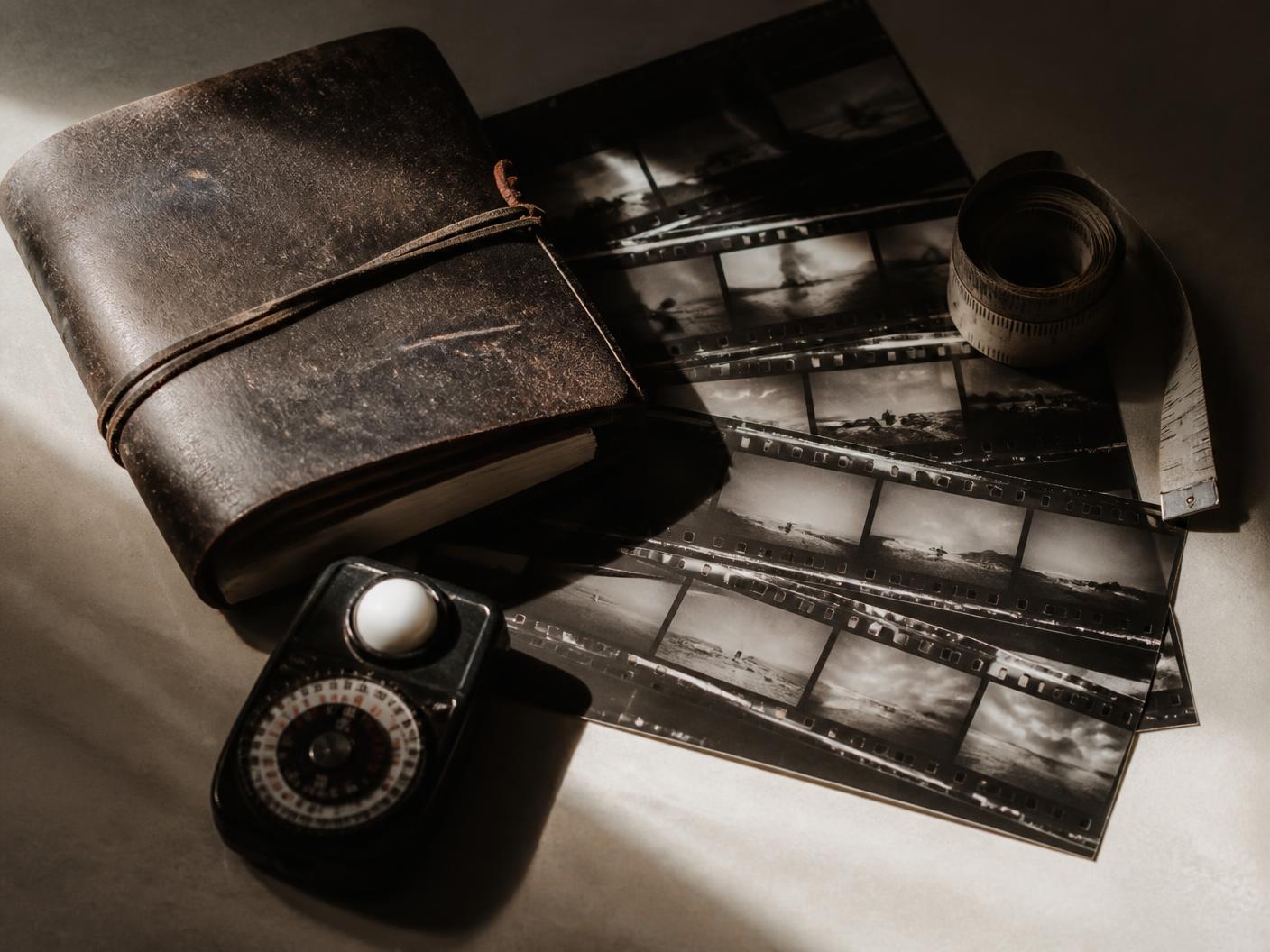 A worn notebook, contact sheets, a light meter and a tape measure on a warm desk
